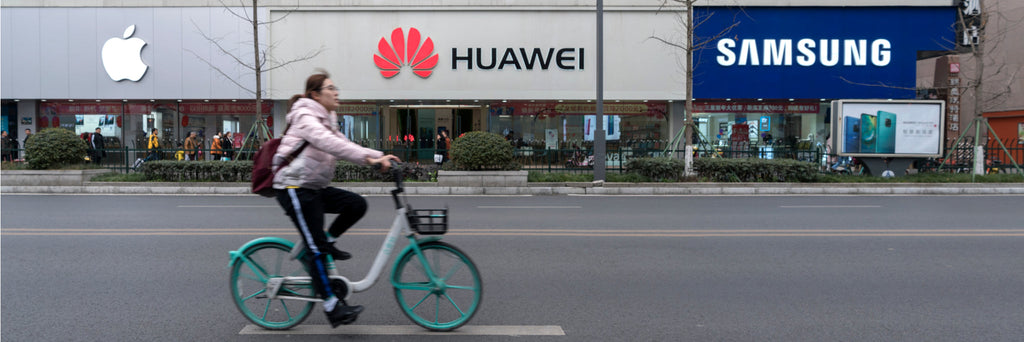 City street view of a person on a bicycle passing Apple, Huawei, and Samsung storefronts, illustrating the wide range of unlocked smartphones that pair perfectly with Gen Mobile’s affordable, reliable high-speed prepaid plans.