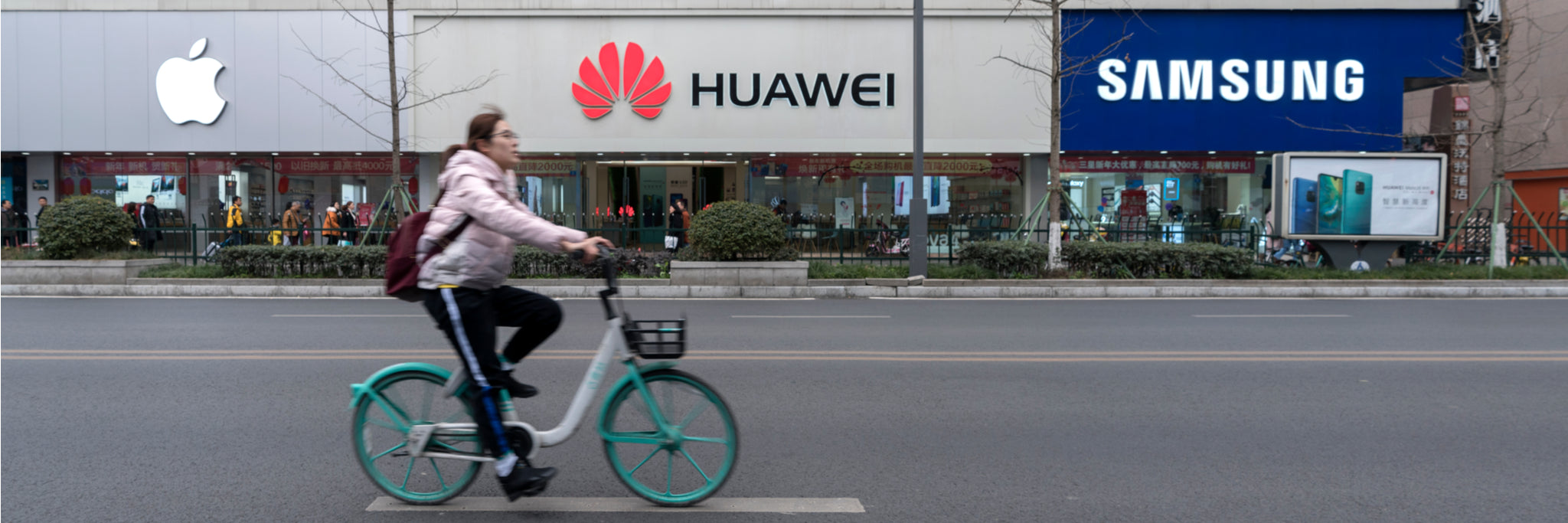 City street view of a person on a bicycle passing Apple, Huawei, and Samsung storefronts, illustrating the wide range of unlocked smartphones that pair perfectly with Gen Mobile’s affordable, reliable high-speed prepaid plans.
